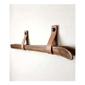 May include: A wall-mounted display rail consisting of a long, curved dark brown wooden bar suspended by two tan leather straps. Each strap is secured to the wall with a small, round black knob, creating a horizontal hanging storage system.