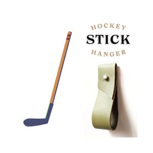 May include: A green leather hockey stick hanger with a silver rivet. The hanger is designed to hold a hockey stick on a wall. The text "HOCKEY STICK HANGER" is printed in black on a white background.