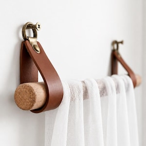 May include: A wall-mounted towel rail featuring a light-coloured wooden rod suspended by two brown leather straps. The straps are secured to the wall with antique brass-toned metal hardware and screws. A sheer white fabric is draped over the wooden rod.
