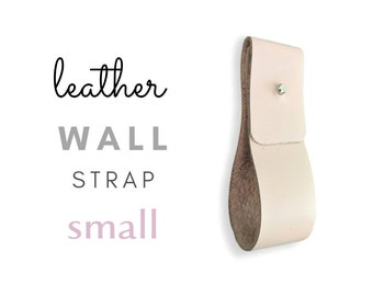 May include: A small, light pink leather wall strap with a silver metal button. The strap is designed to hang items on a wall.