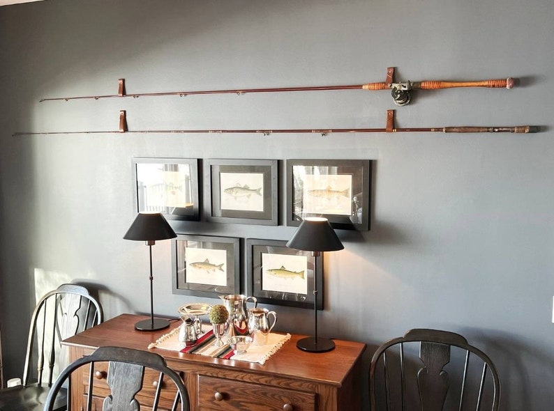 Peut inclure: Deux cannes &agrave; p&ecirc;che en bois mont&eacute;es sur un mur avec des sangles en cuir. Les cannes sont expos&eacute;es au-dessus d'une table en bois avec des lampes noires et des tirages encadr&eacute;s de poissons.