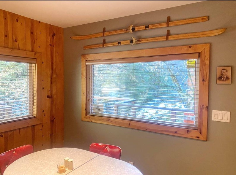 Peut inclure: Une table &agrave; manger en bois avec une surface blanche et deux chaises rouges. La table est &eacute;quip&eacute;e d'un set de sel et de poivre. La pi&egrave;ce a un mur en bois et une fen&ecirc;tre avec des stores. Il y a deux paires de skis accroch&eacute;s au mur.