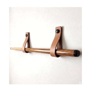 Op de afbeelding: Een wandgemonteerde handdoekhouder bestaande uit een gladde, cilindrische houten stang met zwarte eindkappen, opgehangen aan twee beige lederen riemen. Elke lederen riem is aan de muur bevestigd met een zwarte ronde knop aan de bovenzijde.