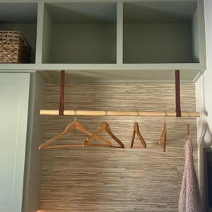 Könnte beinhalten: Eine Holzstange mit braunen Lederriemen, die an einem grünen Regal hängen. Vier Holzbügel hängen an der Stange. Die Wand hinter der Stange ist mit einer hellbraunen, strukturierten Tapete bedeckt.
