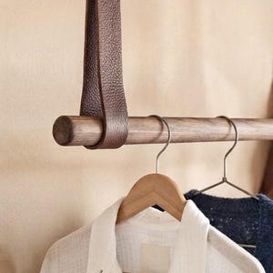 May include: A horizontal wooden rod suspended by a dark brown textured leather strap. Two wooden clothes hangers are hooked onto the rod, holding a white collared shirt and a dark blue knit garment, showcasing a functional clothing storage system.