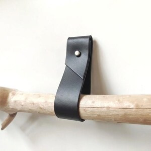 May include: A black leather strap folded into a loop and secured with a single silver-toned metal rivet. The strap is draped over a light-colored, textured wooden branch that extends horizontally across the frame against a plain white background.