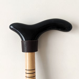Op de afbeelding: Een zwarte houten wandelstok met een gebogen handvat en een bruine leren wikkel. De stok heeft een natuurlijke houtkleur en is versierd met zwarte lijnen.