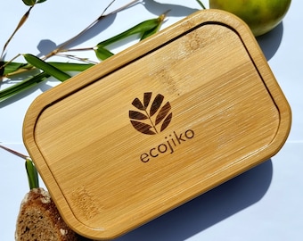Op de afbeelding: Een rechthoekige lunchbox van natuurlijk hout met een afgeronde rand en een logo met de tekst "ecojiko" met een gestileerd bladerenontwerp.