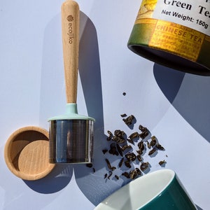 Puede incluir: Un infusor de té de madera con una parte superior de silicona azul claro y un filtro de malla de acero inoxidable. El infusor está sobre una superficie blanca con hojas de té verde sueltas esparcidas a su alrededor. Las hojas de té están en un recipiente cilíndrico verde y amarillo con el texto "Green Tea" y "Chinese Tea" impreso en él. El recipiente tiene un peso neto de 150g.