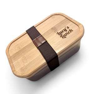 May include: A rectangular bamboo lunch box with a brown elastic strap and a small leather patch. The lid is engraved with the words "Lucy's Lunch" in a cursive font. The box has a light brown wooden lid and a dark brown base.