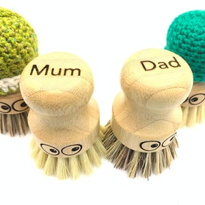 May include: Three wooden dish scrubbers with green crocheted tops. The scrubbers have a round, wooden handle with the words "Mum", "Dad" and a blank space for personalization. Each scrubber has two black eyes and a bristle brush.