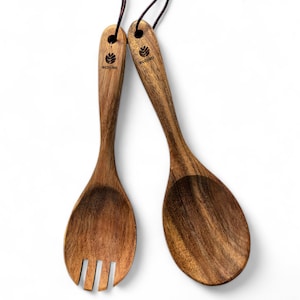 Puede incluir: Un juego de dos utensilios de servicio de madera, una cuchara y un tenedor, con un acabado de grano de madera marrón. Los utensilios están atados juntos con un cordón marrón y tienen la palabra "ecojiko" grabada.