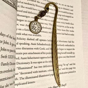 Può includere: Un segnalibro in stile vintage con un ciondolo a forma di orologio e dettagli in metallo ornati. Il segnalibro è color oro con un design curvo e un piccolo quadrante che indica l'ora.