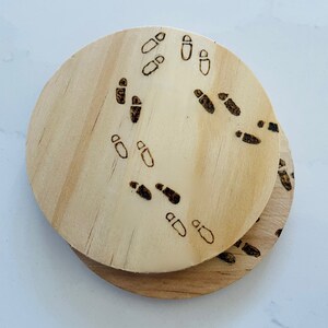 May include: Two round wooden coasters with a burnt design of footprints. The coasters are stacked on top of each other.