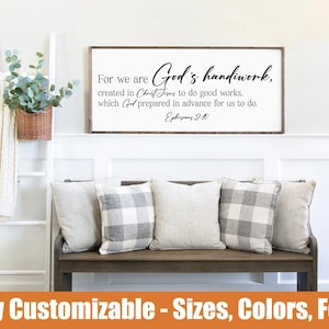 May include: A framed sign with the text "For we are God's handiwork" above a bench with pillows. The sign is in a white frame. The bench has four pillows with a mix of patterns. A decorative ladder and a basket of greenery are on the left.
