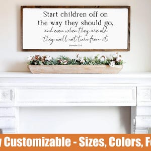 May include: A rectangular wooden framed sign with the text "Start children off on the way they should go, and even when they are old they will not turn from it. Proverbs 22:6". Below the sign is a white mantel with a wooden trough filled with greenery.