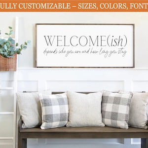 May include: A wooden framed sign with the text "WELCOME(ish) depends who you are and how long you stay". The sign is above a wooden bench with four pillows. A white ladder with a blanket and a basket of greenery are on the left.
