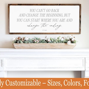 May include: A framed wooden sign with the quote "YOU CAN'T GO BACK AND CHANGE THE BEGINNING, BUT YOU CAN START WHERE YOU ARE AND change the ending." The sign is above a white mantel decorated with a wooden trough filled with greenery and cotton.