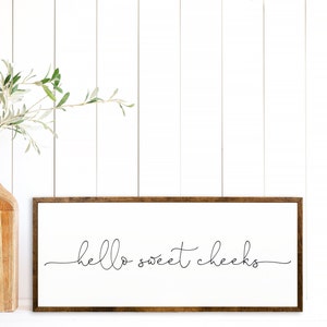 K&ouml;nnte beinhalten: Ein gerahmtes Schild mit dem Text "hello sweet cheeks" in einer einfachen, handgeschriebenen Schrift. Das Schild h&auml;ngt an einer wei&szlig;en Wand mit einer Holzvase und einer gr&uuml;nen Pflanze im Vordergrund.