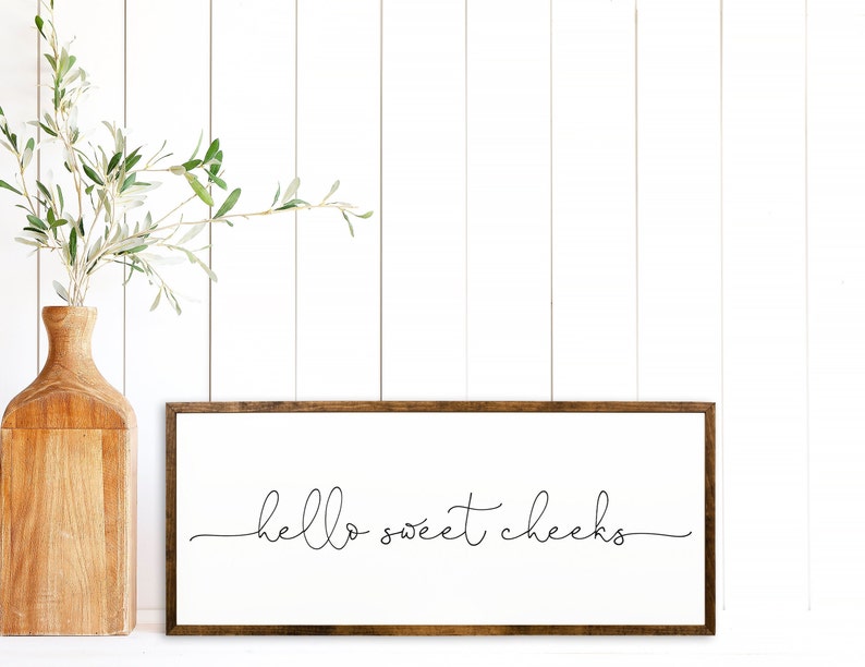 K&ouml;nnte beinhalten: Ein gerahmtes Schild mit dem Text "hello sweet cheeks" in einer einfachen, handgeschriebenen Schrift. Das Schild h&auml;ngt an einer wei&szlig;en Wand mit einer Holzvase und einer gr&uuml;nen Pflanze im Vordergrund.