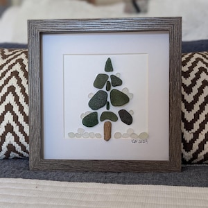 May include: A framed artwork featuring a green glass mosaic tree with a brown wooden trunk. The tree is surrounded by white glass stones, creating a snowy scene. The artwork is signed "KW 2024" in the bottom right corner.