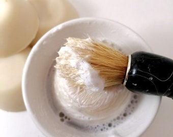 May include: A white shaving soap puck in a white bowl with a shaving brush lathered in white shaving cream.