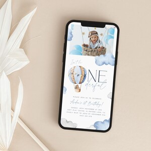 Op de afbeelding: Een digitale uitnodiging voor een eerste verjaardagsfeest met een blauw en wit aquarel-heteluchtballonontwerp. De uitnodiging luidt "I'm the one derful" en bevat de tekst "Please join us to celebrate Andra's 1st Birthday! Sunday, 20th November at 2pm Soft Play Center in Oakwood Lane, CA Kindly RSVP to Josie by November 5th 0412 345 478".