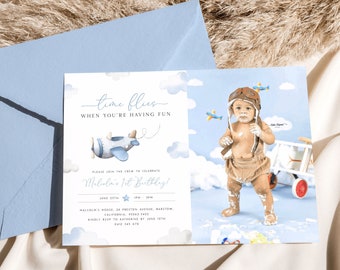 May include: A blue and white airplane themed first birthday party invitation with a photo of a baby boy wearing a pilot's hat and goggles. The invitation says "Time Flies When You're Having Fun" and "Please Join The Crew To Celebrate [Baby's Name]'s 1st Birthday!" with the date and time of the party. The invitation also includes the address and RSVP information.
