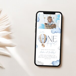 Op de afbeelding: Een digitale uitnodiging in blauw en wit met een aquarel-illustratie van een luchtballon en wolken. De tekst luidt "Isn't he one derful" en "Please join us to celebrate Aden's 1st Birthday!" met details over het evenement.