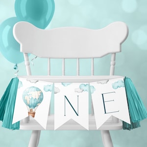 Puede incluir: Una silla alta blanca con una pancarta azul turquesa y blanca que dice "ONE" con un gráfico de globo aerostático. La pancarta tiene borlas azul turquesa en cada extremo.