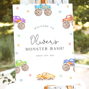 May include: A white welcome sign with a blue, green, orange, and purple monster truck on each corner. The sign says "Welcome to Oliver's Monster Bash! August 12th, 2023".