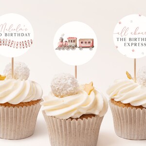 May include: Three cupcakes with white frosting and a white coconut ball on top. Each cupcake has a round paper topper with a train theme. The first topper says "Malcolm's 3rd Birthday" with a pink train track. The second topper has a pink and brown train. The third topper says "All aboard The Birthday Express" with pink stars.