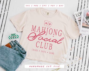 Może przedstawiać: Kremowa koszulka z napisem "MAHJONG Social CLUB" w kolorze różowym. Koszulka posiada grafikę z kafelkami do mahjonga oraz tekst "CRAKS + DOTS + BAMS". Koszulka jest połączona z niebieskimi dżinsami, zielono-białą spinką do włosów i magazynem.