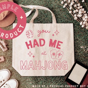 May include: A cream-colored tote bag with the phrase "You Had Me at Mahjong" in pink. The design includes dice, stars, and Mahjong tiles. The bag has a simple design with two handles and is set against a green background.