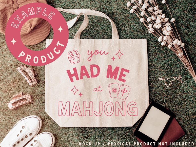 May include: A cream-colored tote bag with the phrase "You Had Me at Mahjong" in pink. The design includes dice, stars, and Mahjong tiles. The bag has a simple design with two handles and is set against a green background.