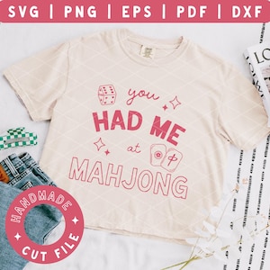 May include: A cream-colored t-shirt with the phrase "you HAD ME at MAHJONG" in pink. The shirt is paired with denim shorts, a green and white hair clip, and a "HANDMADE CUT FILE" sticker. A magazine and flowers are in the background.