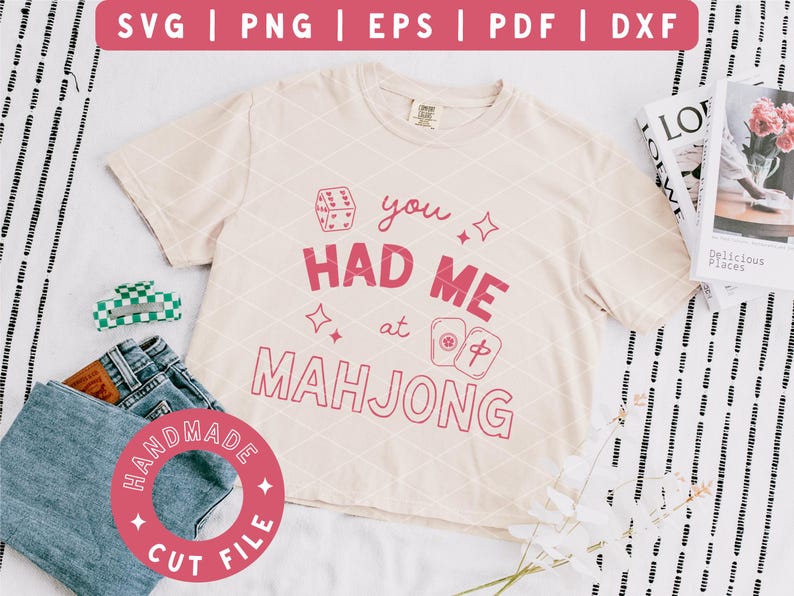 May include: A cream-colored t-shirt with the phrase "you HAD ME at MAHJONG" in pink. The shirt is paired with denim shorts, a green and white hair clip, and a "HANDMADE CUT FILE" sticker. A magazine and flowers are in the background.