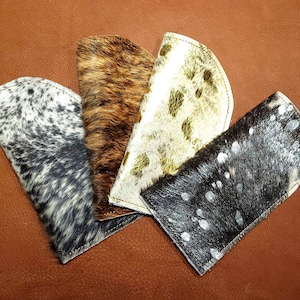 May include: Four different cowhide eyeglass cases in various patterns. The cases are all made of leather and have a soft, suede-like texture. The patterns include black and white, brown and white, and gold and white.