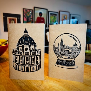 May include: Two brown paper notebooks with black ink illustrations. The notebook on the left features a black and white illustration of a building with a dome. The notebook on the right features a black and white illustration of a snow globe with a cityscape inside. The snow globe has the word "OXFORD" printed on the base.