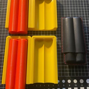 Könnte beinhalten: Zwei gelbe Kunststoffformen zum Herstellen von Zwei-Zigarren-Etuis und ein schwarzes Lederetui mit roten Nähten.