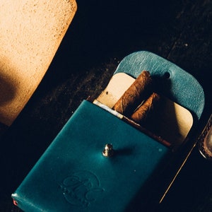 Op de afbeelding: Een teal leren sigarenkoker met een drukknoop sluiting en een rond logo met de tekst "Clay & Milk" in een gestileerd lettertype. Twee bruine sigaren bevinden zich in de koker.