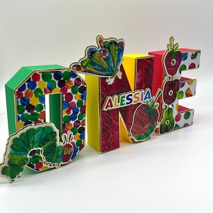 May include: Colorful cardboard letters spelling "ONE" with a green caterpillar, a butterfly, and strawberries. The letter "N" has the name "ALESSIA" written on it.
