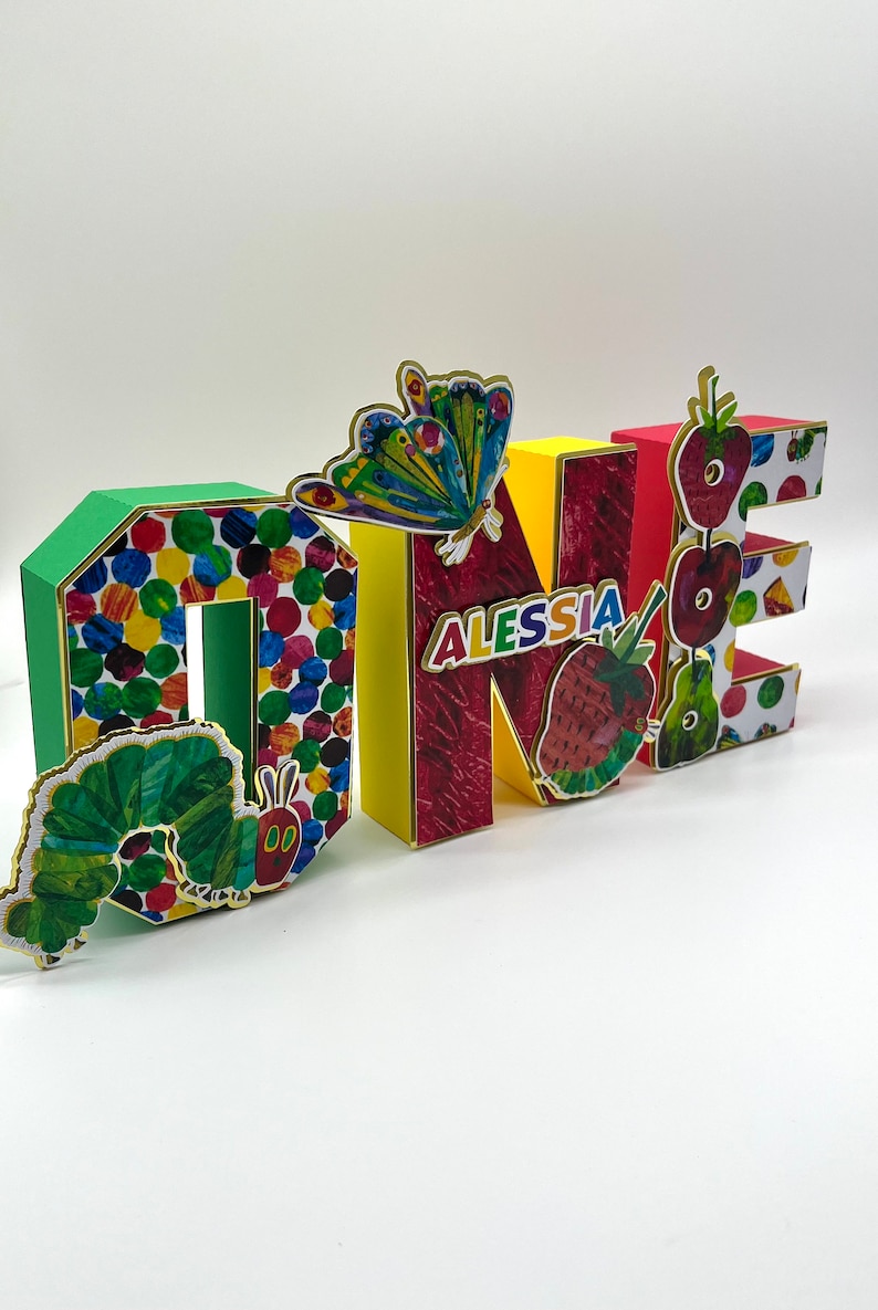 May include: Colorful cardboard letters spelling "ONE" with a green caterpillar, a butterfly, and strawberries. The letter "N" has the name "ALESSIA" written on it.