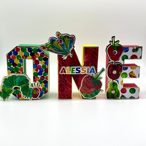 May include: Colorful wooden letters spelling "ONE" with a butterfly, strawberries, and a caterpillar from the book "The Very Hungry Caterpillar" on them. The letters are decorated with colorful dots and the name "ALESSIA" is written on the "N".