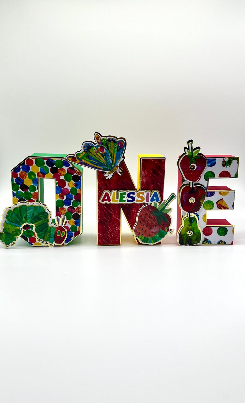 May include: Colorful wooden letters spelling "ONE" with a butterfly, strawberries, and a caterpillar from the book "The Very Hungry Caterpillar" on them. The letters are decorated with colorful dots and the name "ALESSIA" is written on the "N".