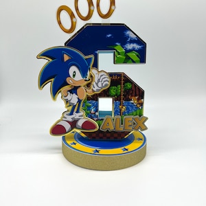 May include: A blue and gold Sonic the Hedgehog themed cake topper with the number 6, the character Sonic, and the name Alex. The topper is decorated with gold rings and stars.