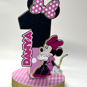 May include: Pink and black Minnie Mouse themed centerpiece with a large number one, the name "Danya" and a Minnie Mouse figure. The base is pink with white polka dots and a gold glitter trim.