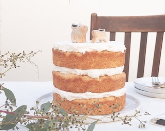 May include: A three-tiered white cake with vanilla frosting, decorated with two small wooden bunny figurines. The cake is on a white plate and there is a white tablecloth underneath. There are pink and white striped flags hanging in the background that say "Olive".