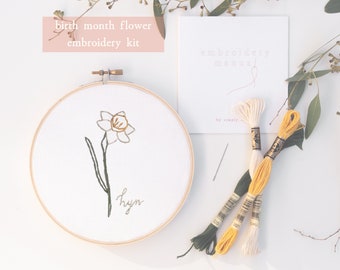 May include: A DIY embroidery kit featuring a white daffodil flower with green leaves and the word "hyacinth" embroidered on white fabric in a wooden embroidery hoop. The kit includes three skeins of embroidery floss in yellow, cream, and green, a needle, and a card with the words "embroidery kit" printed on it.
