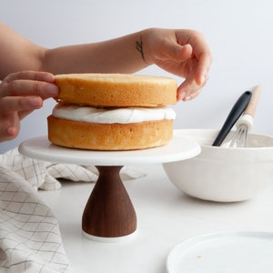 May include: A two-tier cake on a white cake stand with a wooden base. The cake is made of two yellow cake layers and is frosted with white frosting.